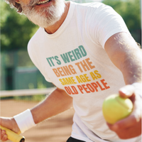 White tee featuring bold, rainbow-colored lettering reads It’s Weird Being the Same Age as Old People 