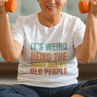 White tee featuring bold, rainbow-colored lettering reads It’s Weird Being the Same Age as Old People 