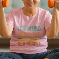 Pink tee featuring bold, rainbow-colored lettering reads It’s Weird Being the Same Age as Old People 