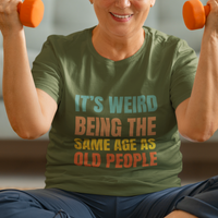 Military green tee featuring bold, rainbow-colored lettering reads It’s Weird Being the Same Age as Old People 
