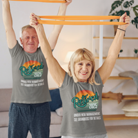 A couple in gray t-shirts featuring a rainbow striped sun that reads Retired 2025 Under New Management See Grandkids for Details.