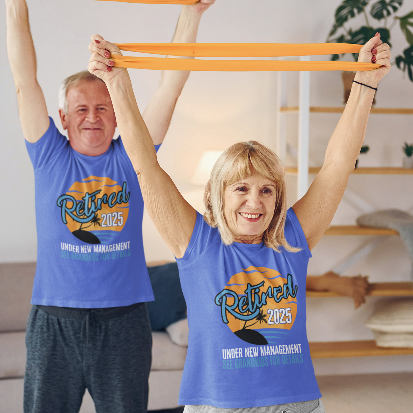 A couple in royal blue t-shirts featuring a sunset beach scene that reads Retired 2025 Under New Management See Grandkids for Details.