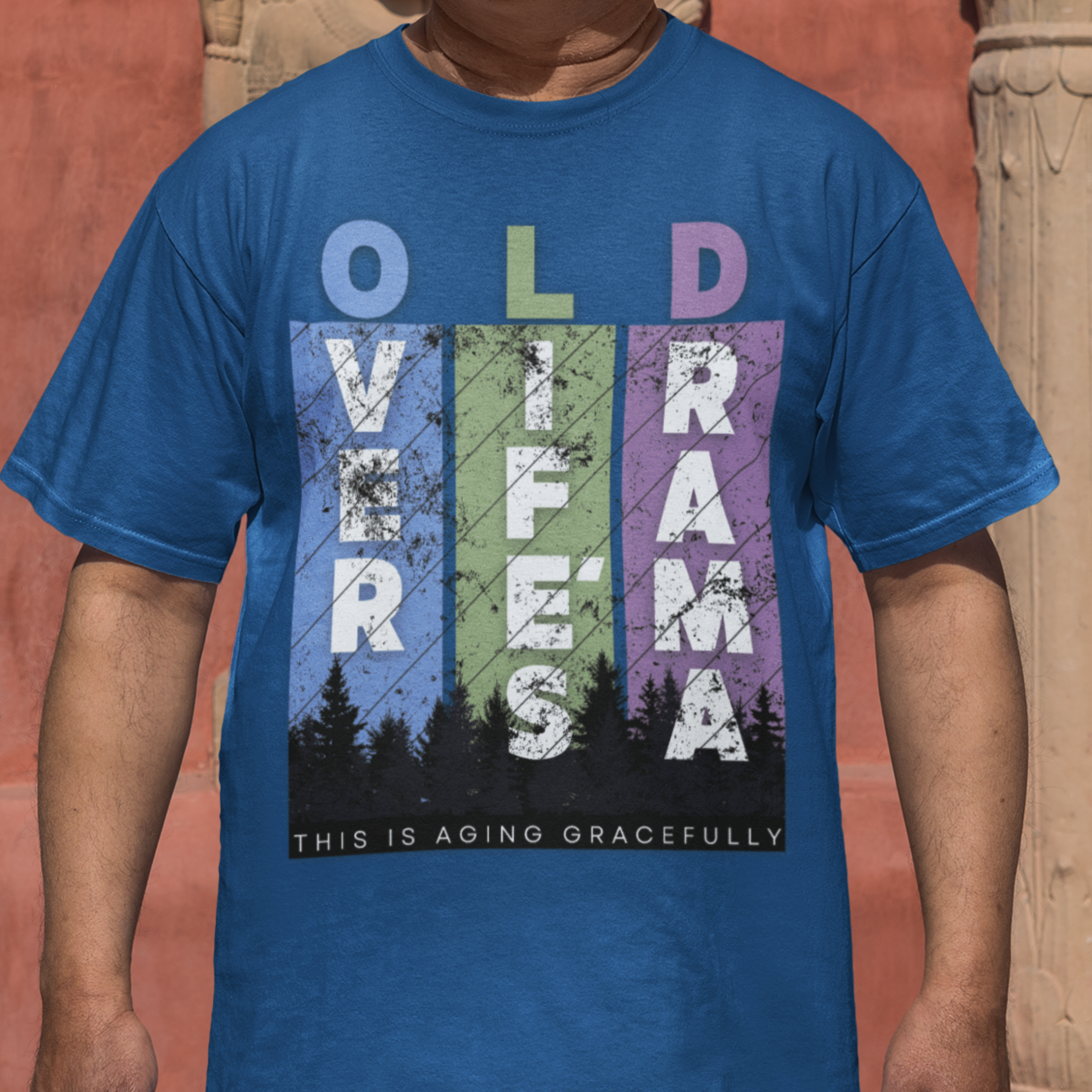 Royal blue t-shirt featuring pastel stripes and lettering with a forest scene in silhouette, stating Over Life's Drama This is Aging Gracefully