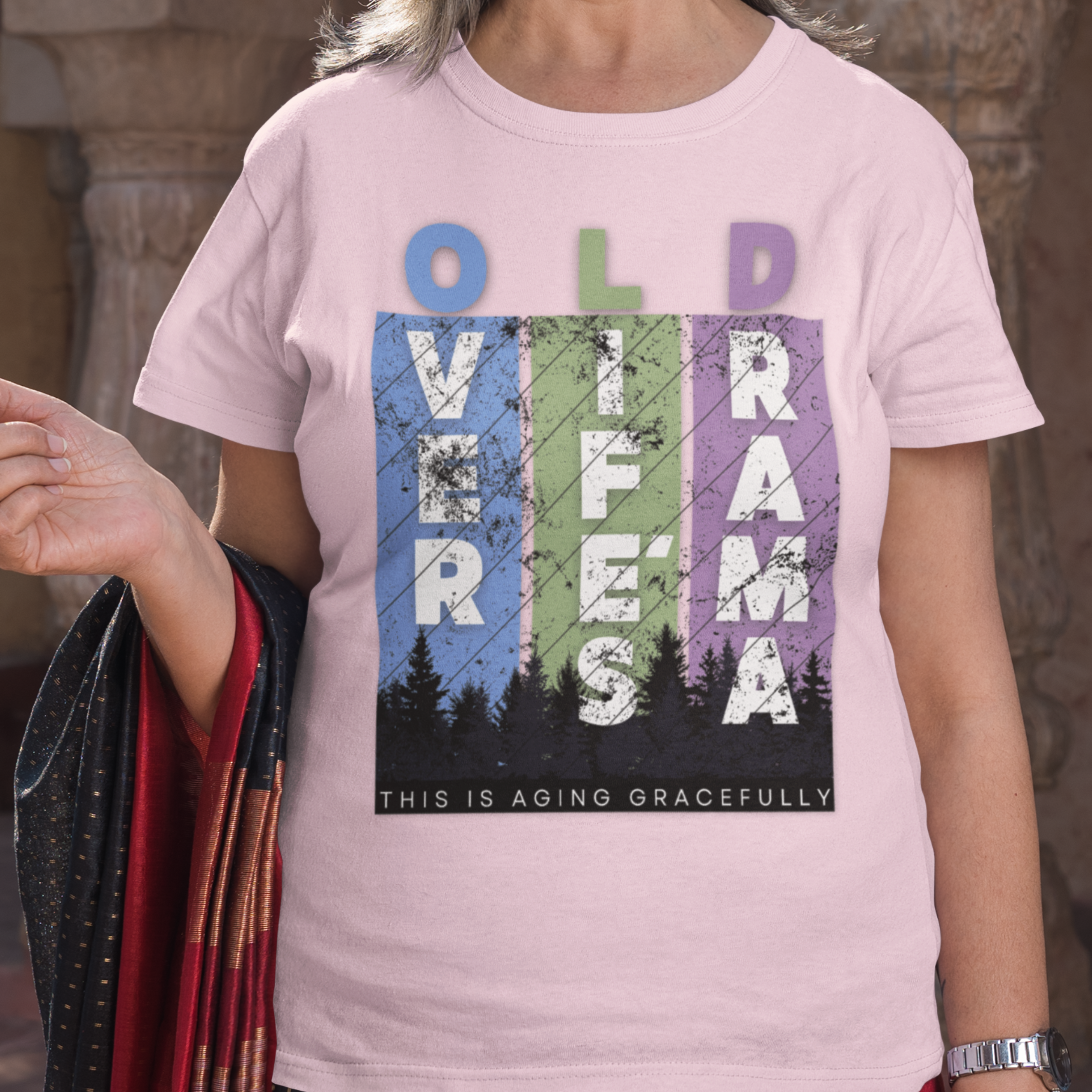 Light pink t-shirt featuring pastel stripes and lettering with a forest scene in silhouette, stating Over Life's Drama This is Aging Gracefully