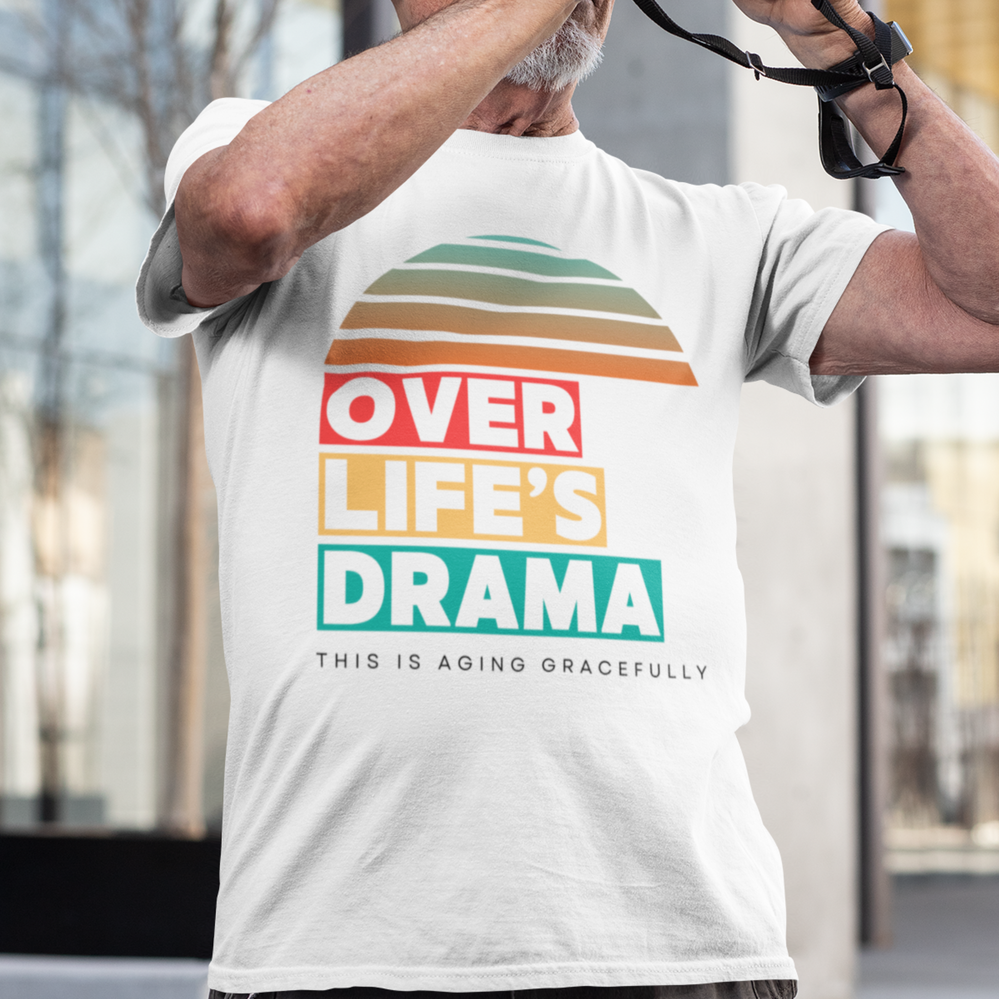White t-shirt that reads Over Life's Drama This is Aging Gracefully, featuring colorful block lettering and a rainbow striped sunset.