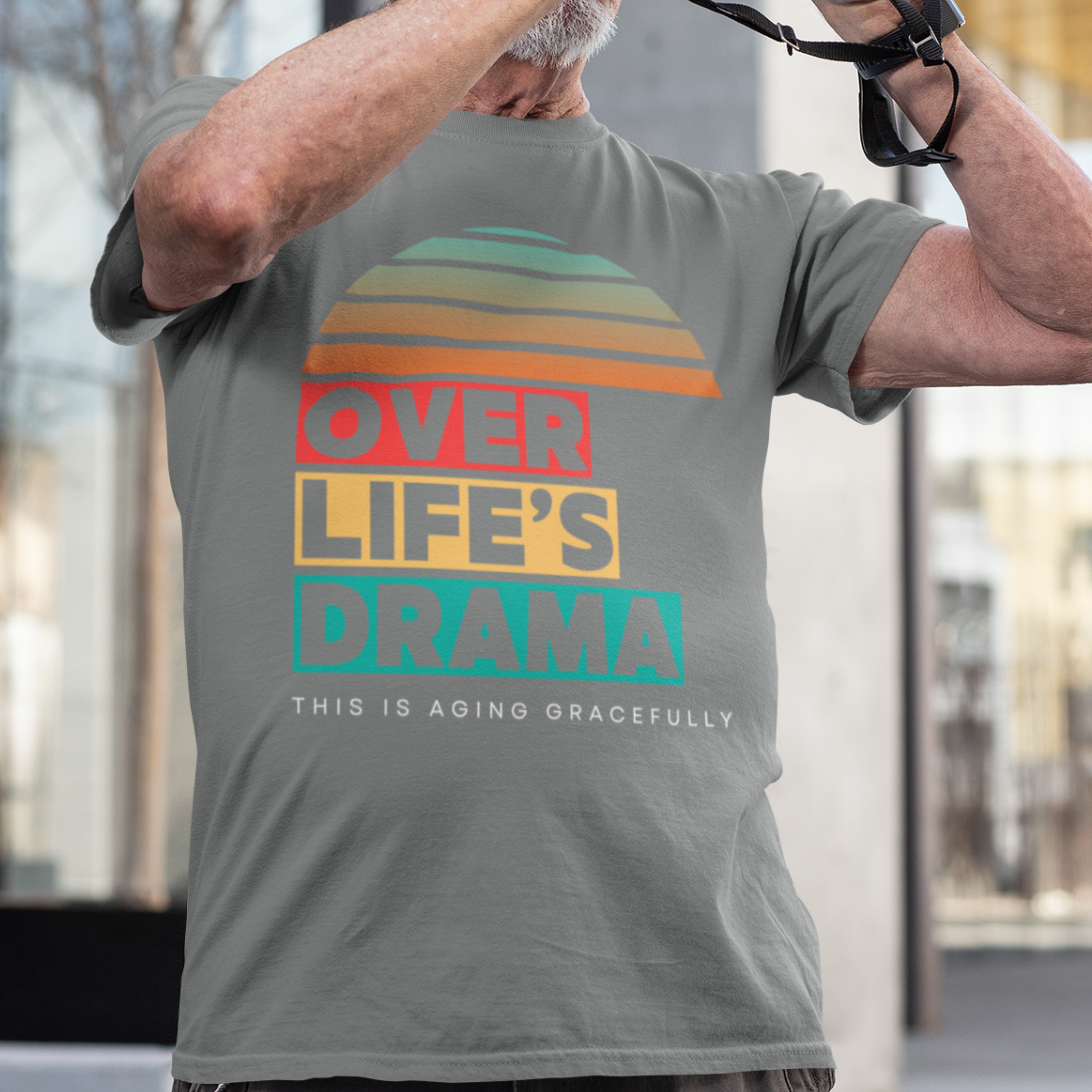 Gray t-shirt that reads Over Life's Drama This is Aging Gracefully, featuring colorful block lettering and a rainbow striped sunset.