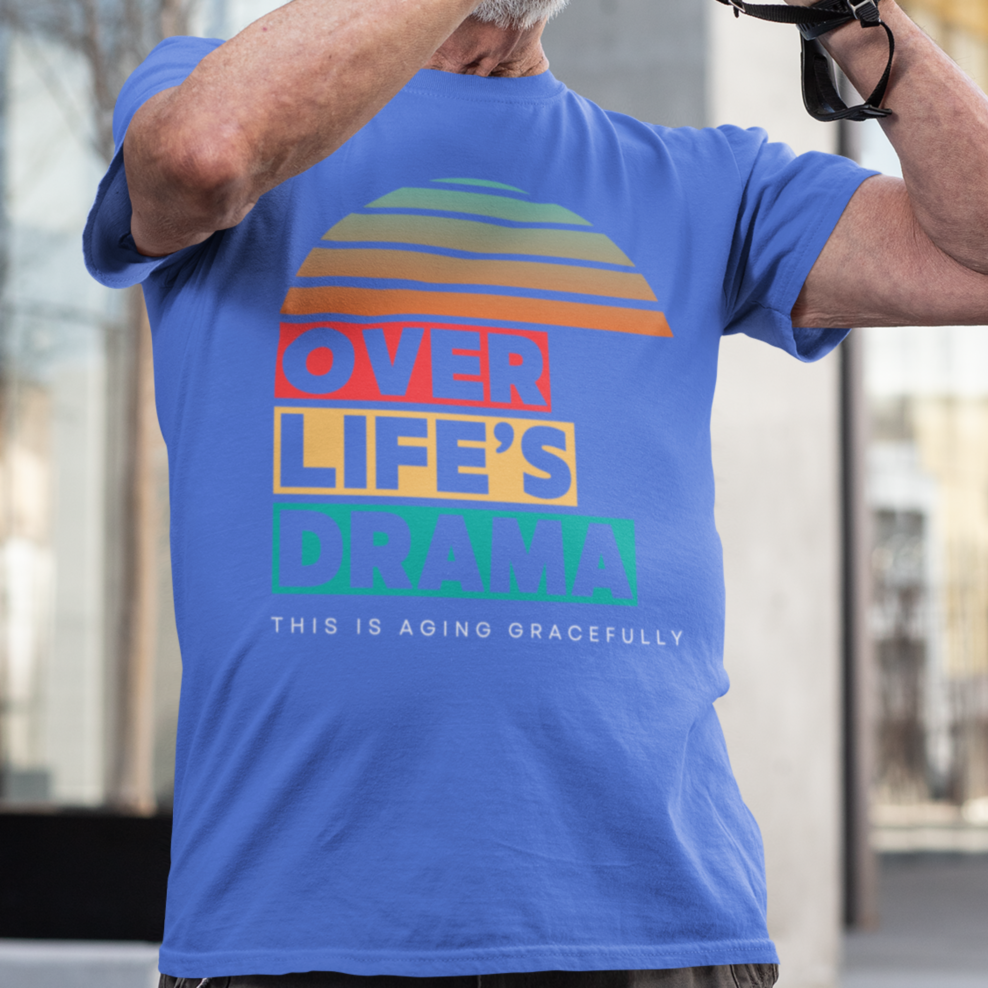 royal blue t-shirt that reads Over Life's Drama This is Aging Gracefully, featuring colorful block lettering and a rainbow striped sunset.