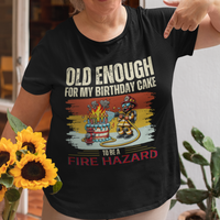 Black tee featuring a cartoon fireman putting out the flames from the candles on a birthday cake.