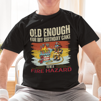 Black tee featuring a cartoon fireman putting out the flames from the candles on a birthday cake.