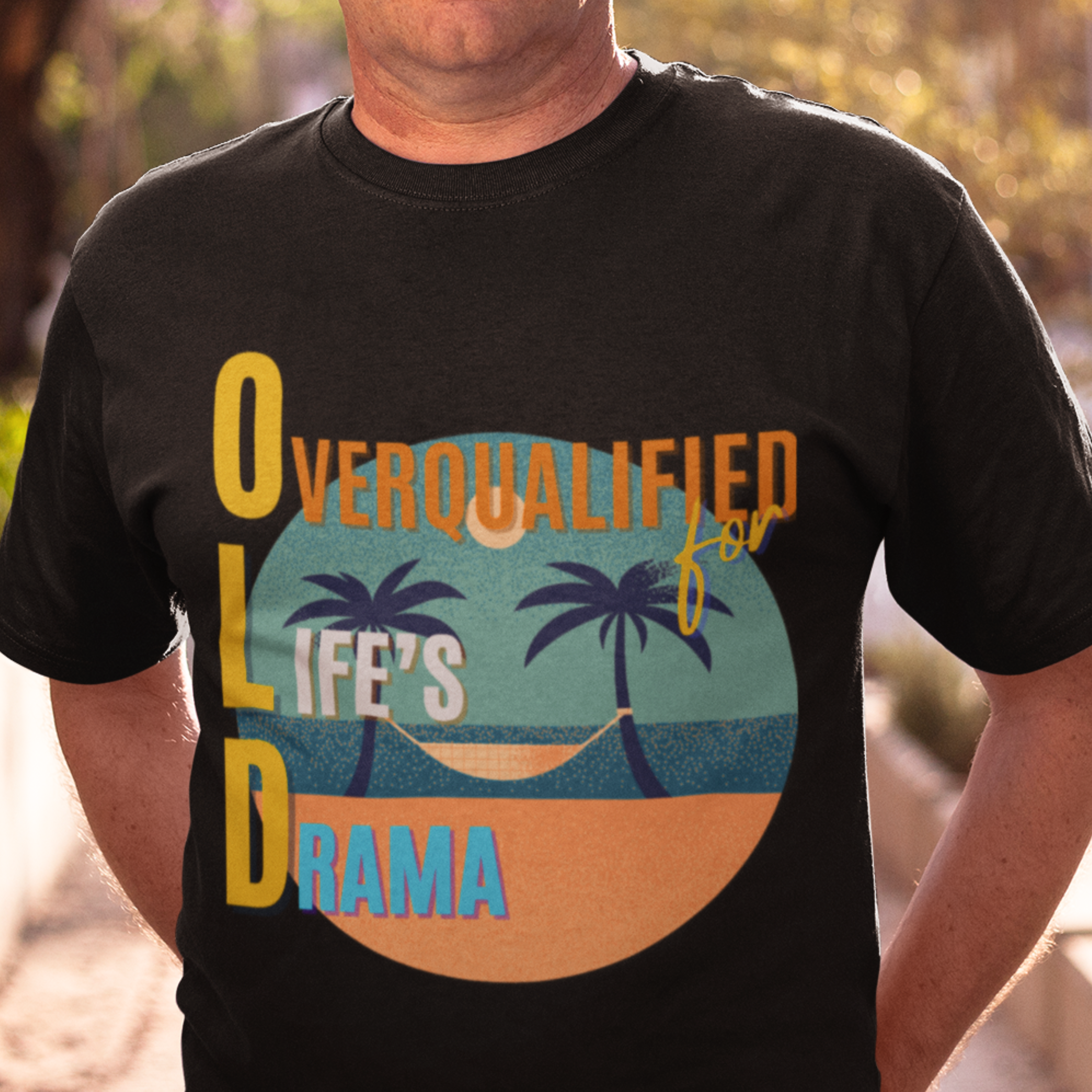 Black tee featuring a hammock between two palm trees on a secluded beach, reading "OLD: Overqualified for Life's Drama"